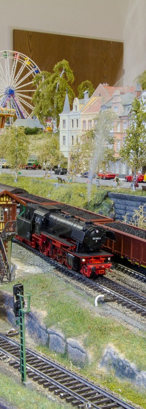 H0-Dampflok fährt durch eine Modelllandschaft mit Karussell im Hintergrund.